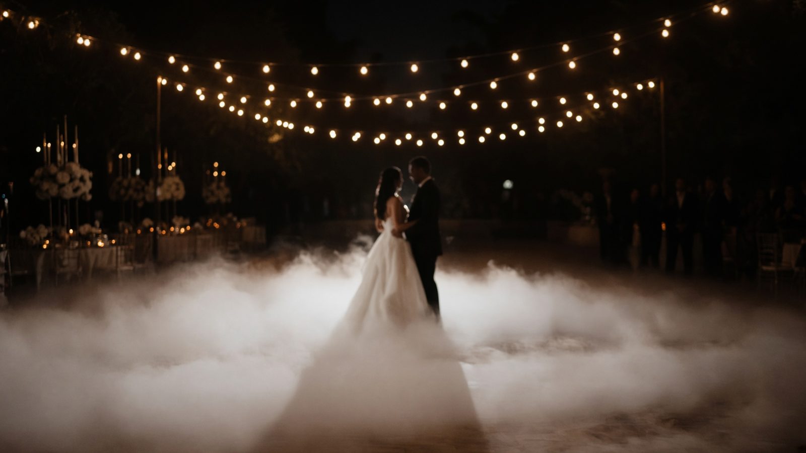 A couple dancing on a reception floor covered in low white fog under string lights.