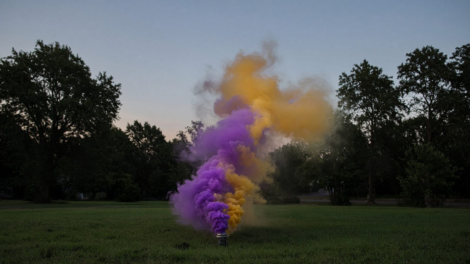 A ground device on a lawn releasing a plume of purple and gold smoke at dusk.