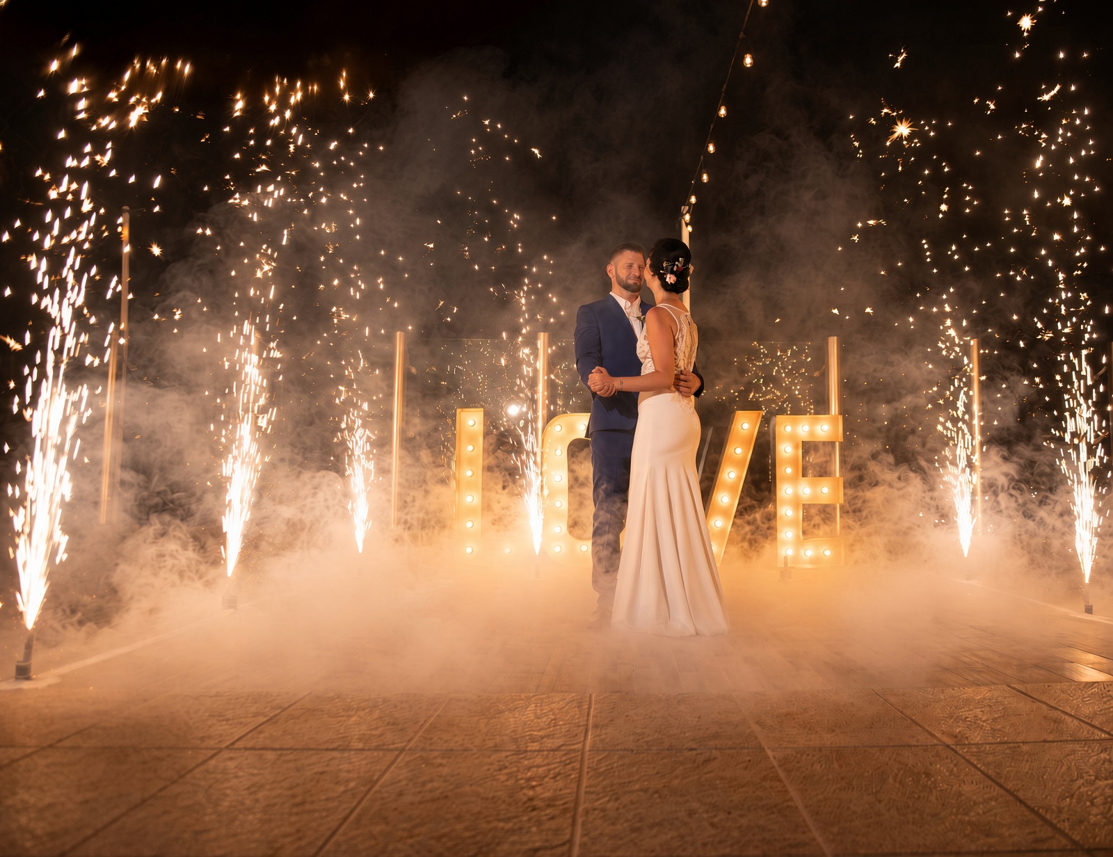 A bride and groom on a night dance floor, with cold-spark fountains, low fog, and a lighted LOVE sign.